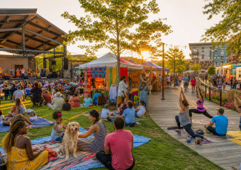 Outdoor festival with live music, market stalls, food trucks, and people doing yoga on a boardwalk